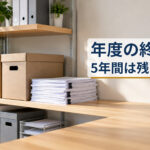 事務所の棚に並んだ書類の箱と、横に置かれた紙の束