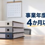 事務所の机に積まれた書類のファイルと、壁のカレンダー