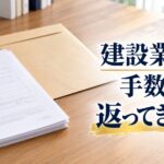 事務所の机に置かれた申請書類の束と、未記入の角形封筒